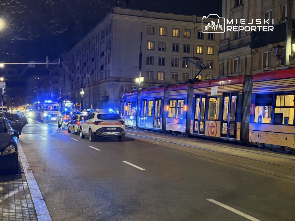 Tramwaj z włączonymi światłami awaryjnymi zatrzymany na ulicy, obok policyjne i pogotowia ratunkowe z niebieskimi sygnałami świetlnymi.