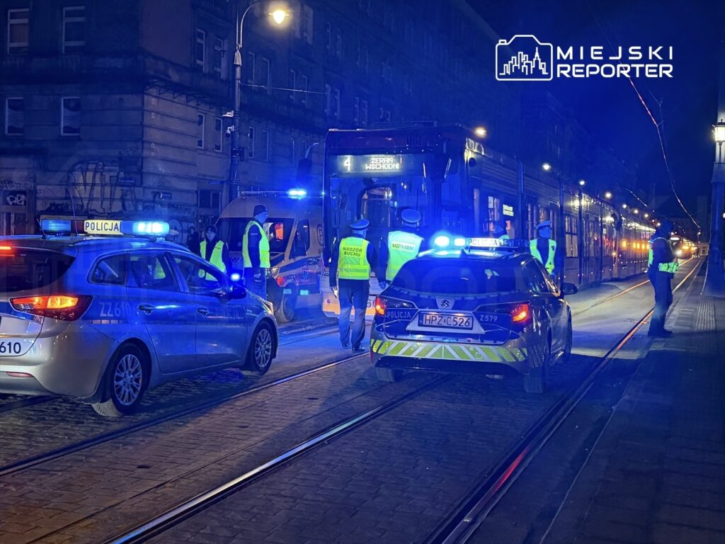 Grupa policjantów w kamizelkach odblaskowych kontroluje miejsce zdarzenia nocą, obok zatrzymanego tramwaju i radiowozów.