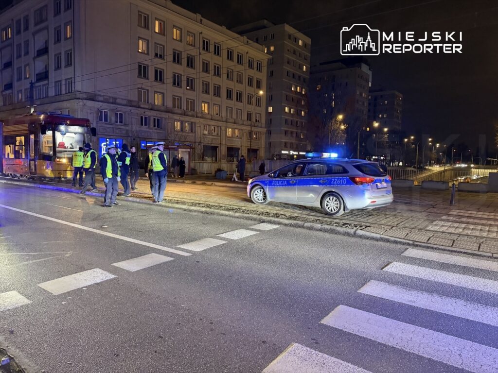 Grupa policjantów w odblaskowych kamizelkach prowadzi działania na ulicy nocą, obok tramwaju i budynków mieszkalnych.