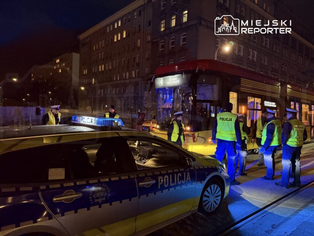 Grupa policjantów w odblaskowych kamizelkach kontroluje tramwaj w nocy, obok stoi radiowóz.