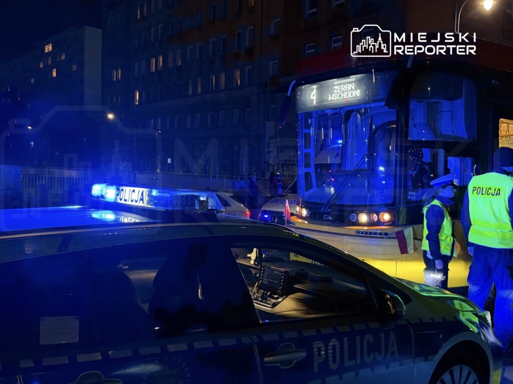 Policjanci w odblaskowych kamizelkach rozmawiają obok autobusu miejskiego z numerem 4, na tle oświetlonych budynków.