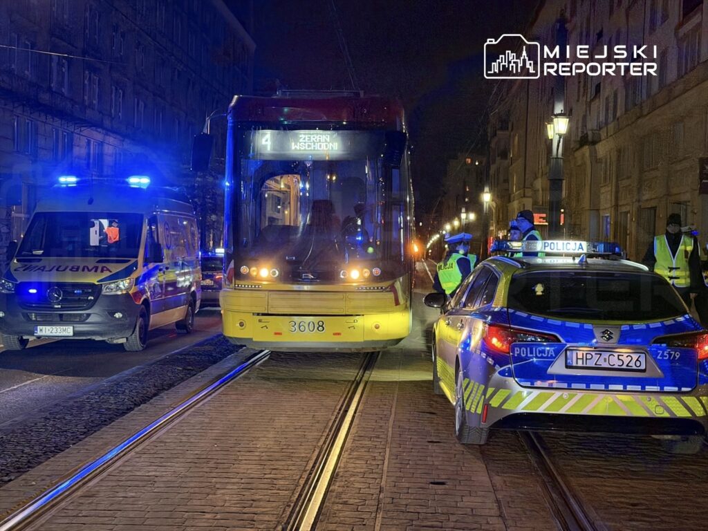 Ambulans i radiowóz policyjny stoją obok tramwaju na ulicy nocą, z włączonymi sygnałami świetlnymi.