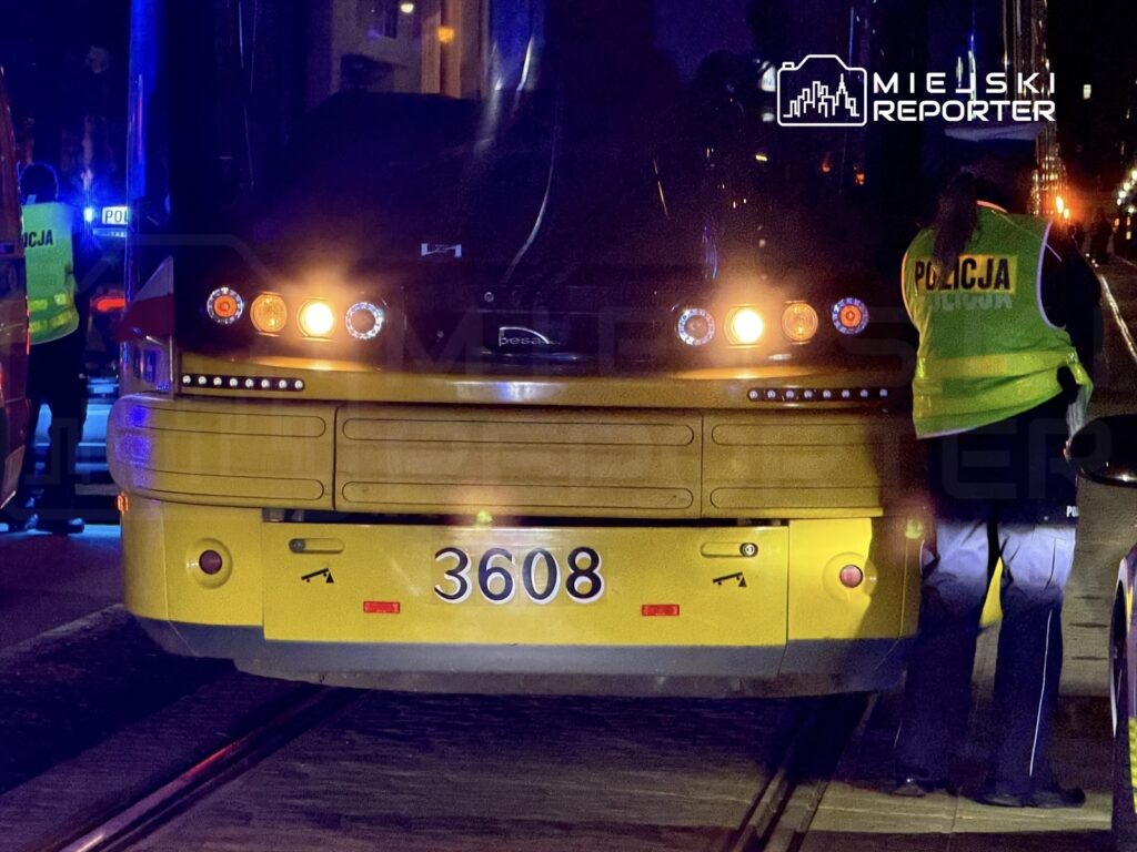Policjant w odblaskowej kamizelce kontroluje żółty tramwaj na torach w nocnym otoczeniu.