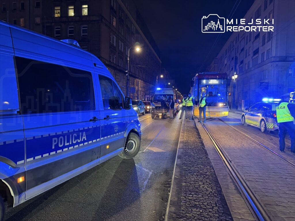 Policyjny radiowóz oraz furgonetka stoją na oświetlonej ulicy, obok zatrzymany tramwaj i funkcjonariusze w kamizelkach odblaskowych.