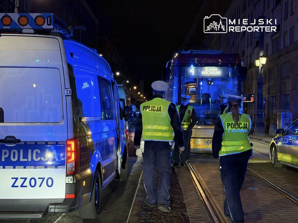Grupa policjantów w kamizelkach odblaskowych kontroluje tramwaj w nocy, otoczona niebieskimi światłami radiowozów.