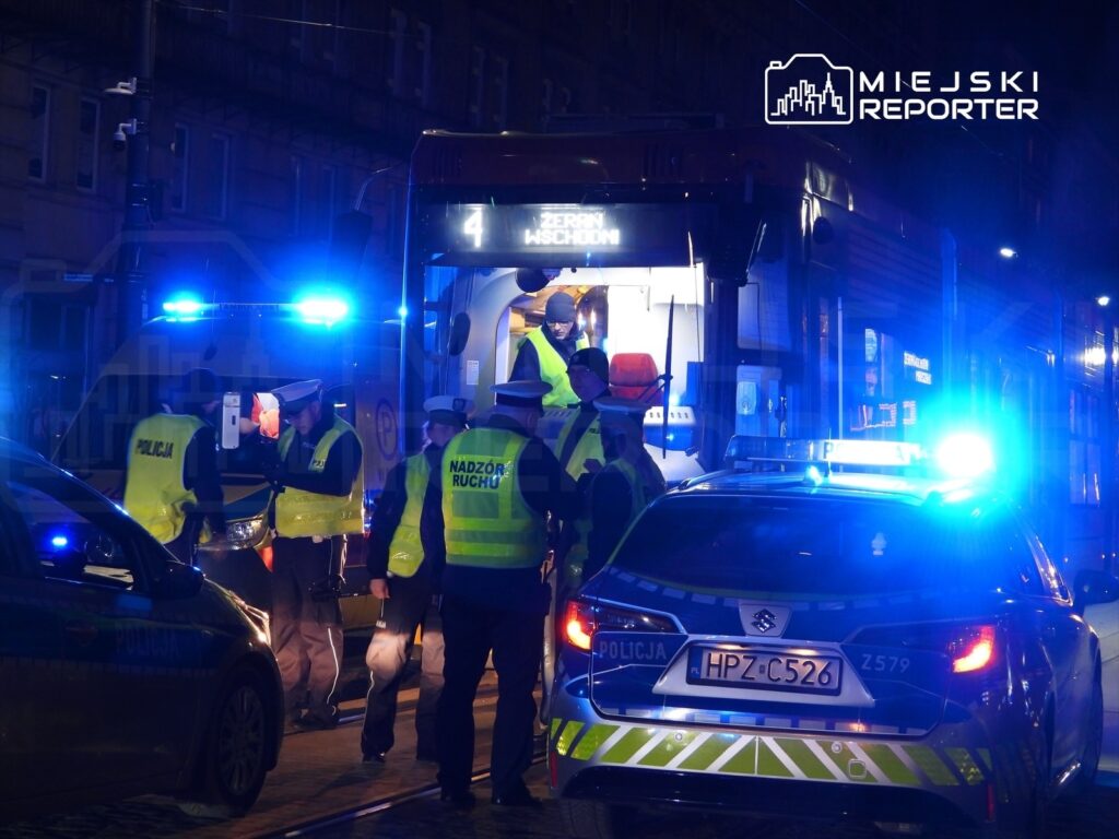 Grupa funkcjonariuszy w kamizelkach odblaskowych kontroluje pasażerów w tramwaju w nocy, otoczeni pojazdami policyjnymi.