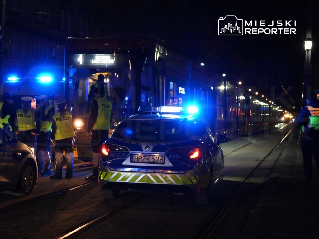 Grupa policjantów w odblaskowych kamizelkach kontroluje pasażerów tramwaju na nocnej ulicy. Obok stoi radiowóz z włączonymi sygnałami świetlnymi.