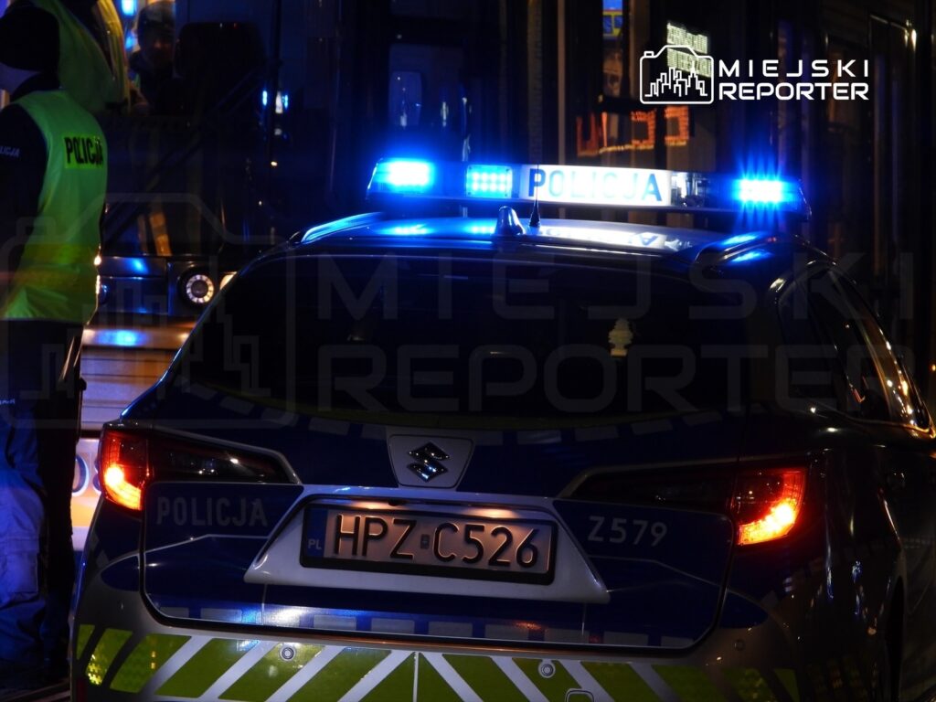 Policyjny samochód z niebieskimi światłami staje obok przystanku, gdzie policjanci w kamizelkach rozmawiają z pasażerami.