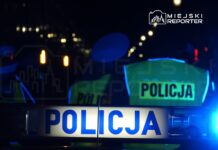 Policjanci w odblaskowych kamizelkach stoją przy radiowozie na oświetlonej ulicy nocą.