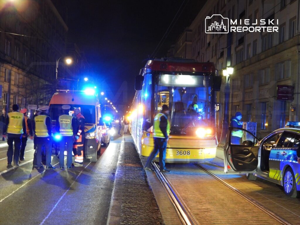 Grupa ratowników i policjantów prowadzi działania na ulicy, obok zatrzymanego tramwaju i karetki.