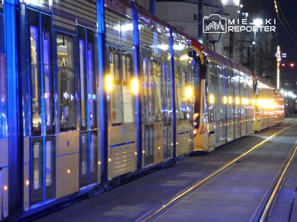 Tramwaj oświetlony niebieskimi i żółtymi światłami zatrzymany na stacji w nocy.