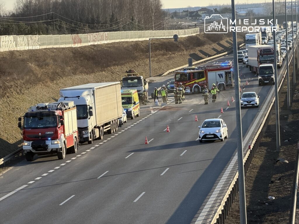 Zespół ratunkowy pracuje przy wypadku drogowym na pasie ruchu, otoczony przez pojazdy i pachołki ostrzegawcze.