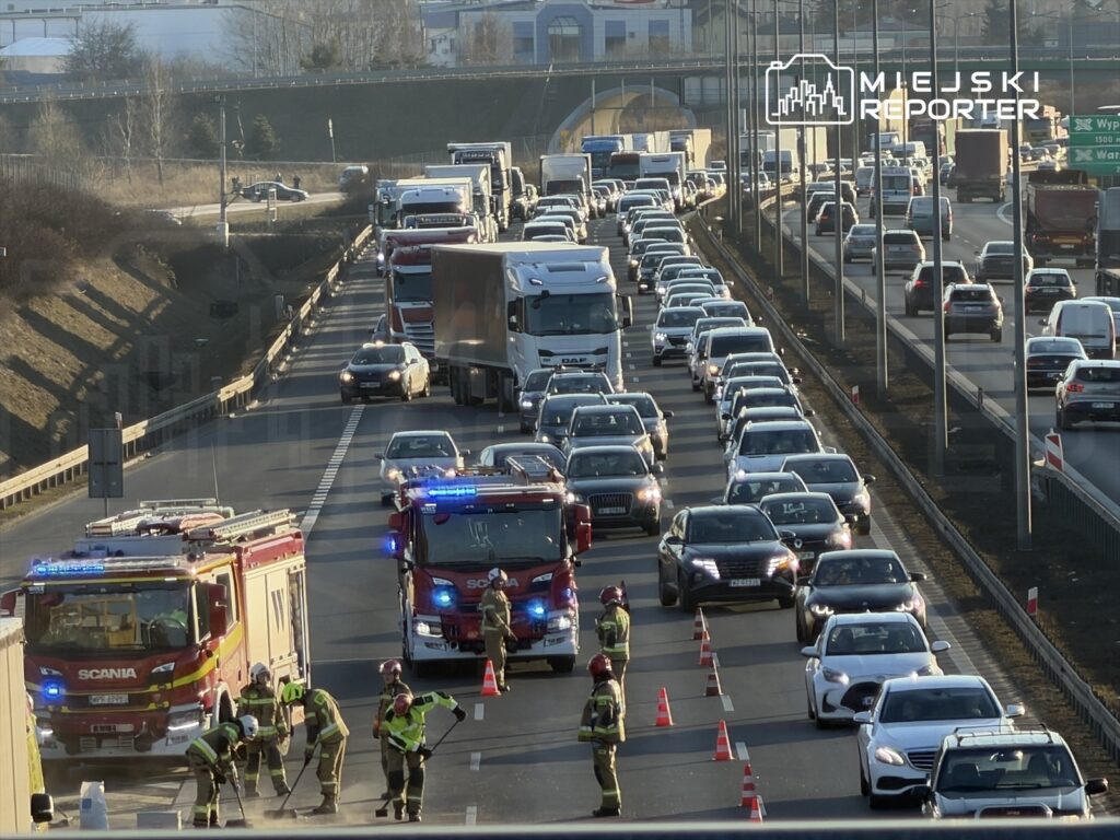 Cztery pojazdy straży pożarnej oraz kilka osób w mundurach pracuje przy rozkopanej jezdni wśród zatoru drogowego.