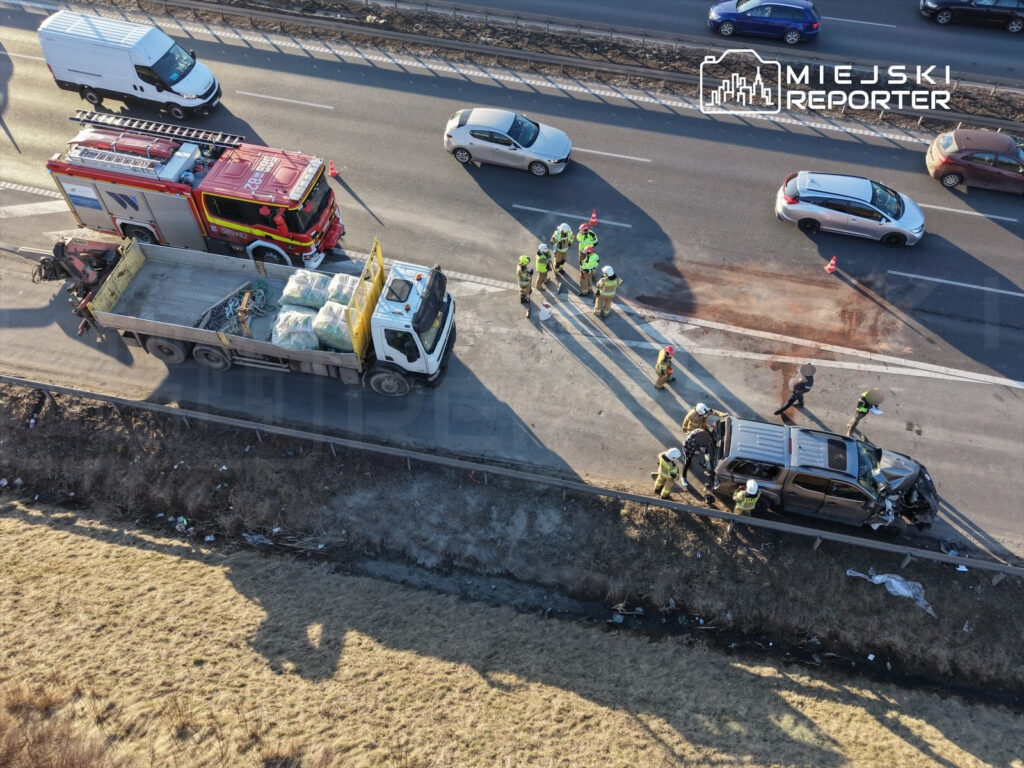 Zespół ratunkowy pracuje przy wypadku drogowym na autostradzie, obok uszkodzonego pojazdu i straży pożarnej.