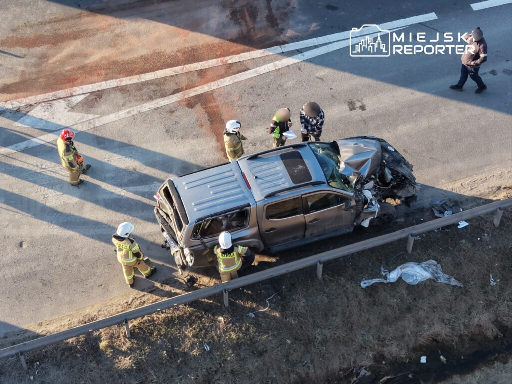 Grupa strażaków w ubraniach ochronnych ocenia uszkodzony samochód po wypadku drogowym na poboczu.