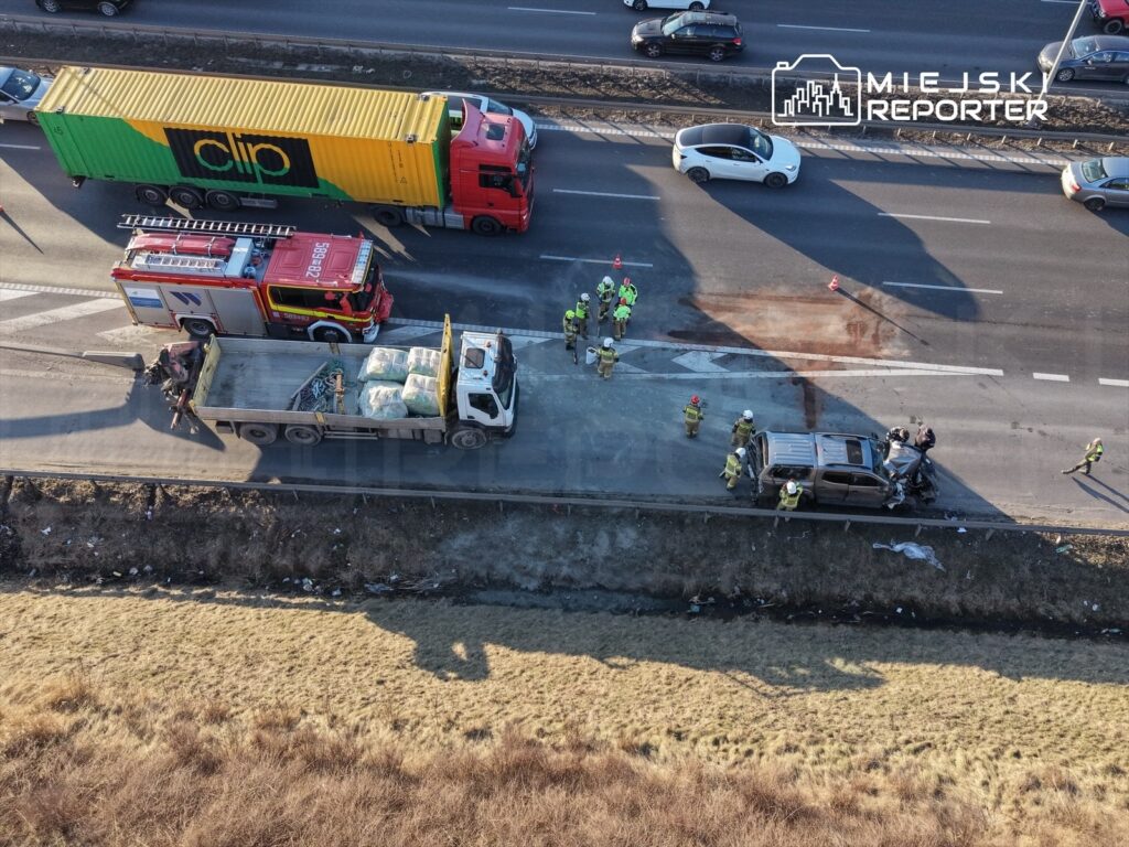 Grupa ratowników i strażaków pracuje przy rozbitym samochodzie na poboczu drogi, obok ciężarówek.