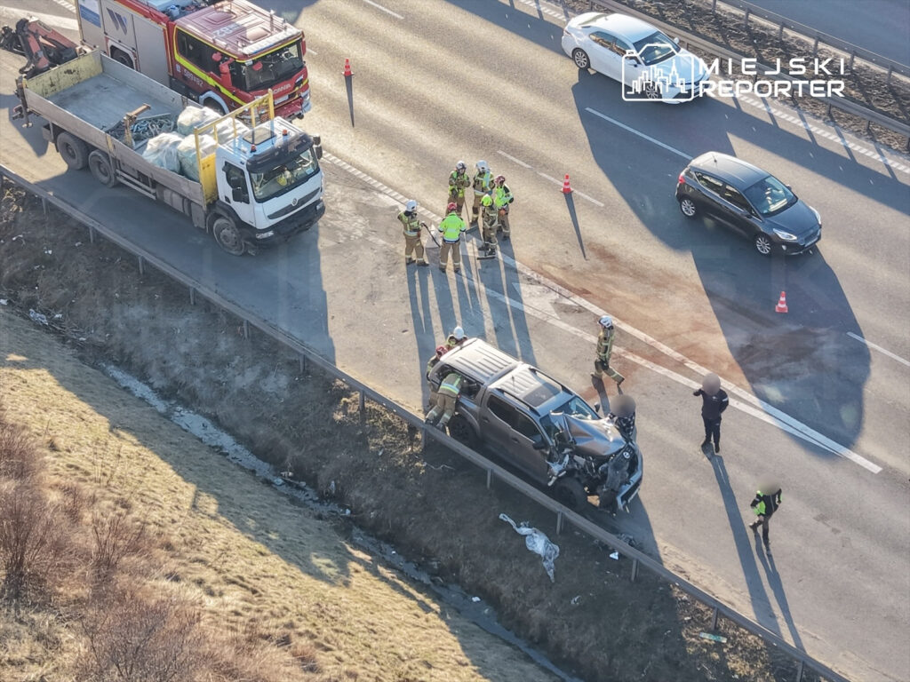 Grupa ratowników i policjantów pracuje przy wypadku drogowym na poboczu, obok uszkodzonego pojazdu.