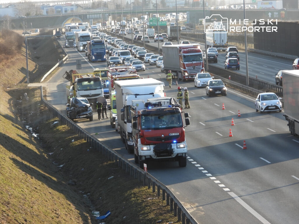 Wielopojazdowy wypadek na autostradzie, w którym biorą udział strażacy oraz pojazdy ratunkowe, z ustawionymi pachołkami.