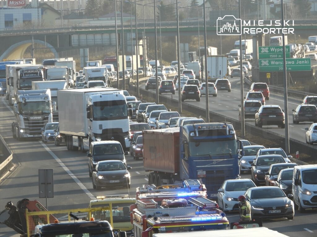 Korki na drodze ekspresowej, z dużą ilością samochodów osobowych i ciężarowych, w tle widoczny most.