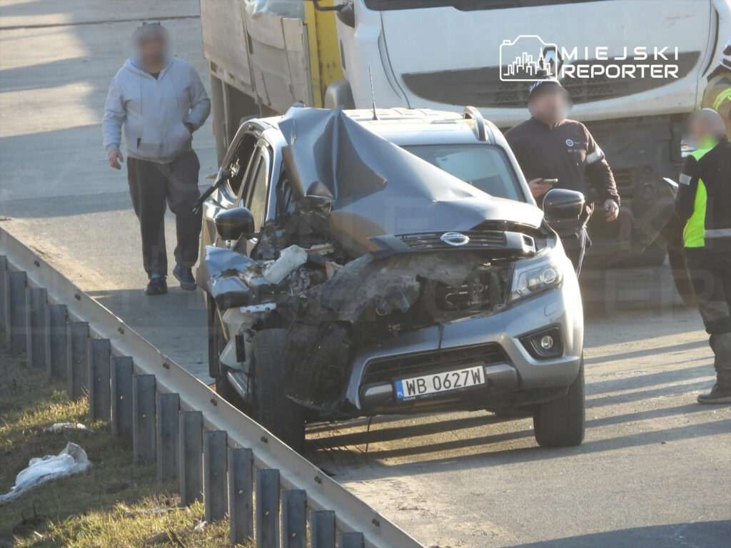 Mężczyzna w szarej bluzie idzie obok uszkodzonego samochodu na drodze, obok stoją pracownicy służb drogowych.