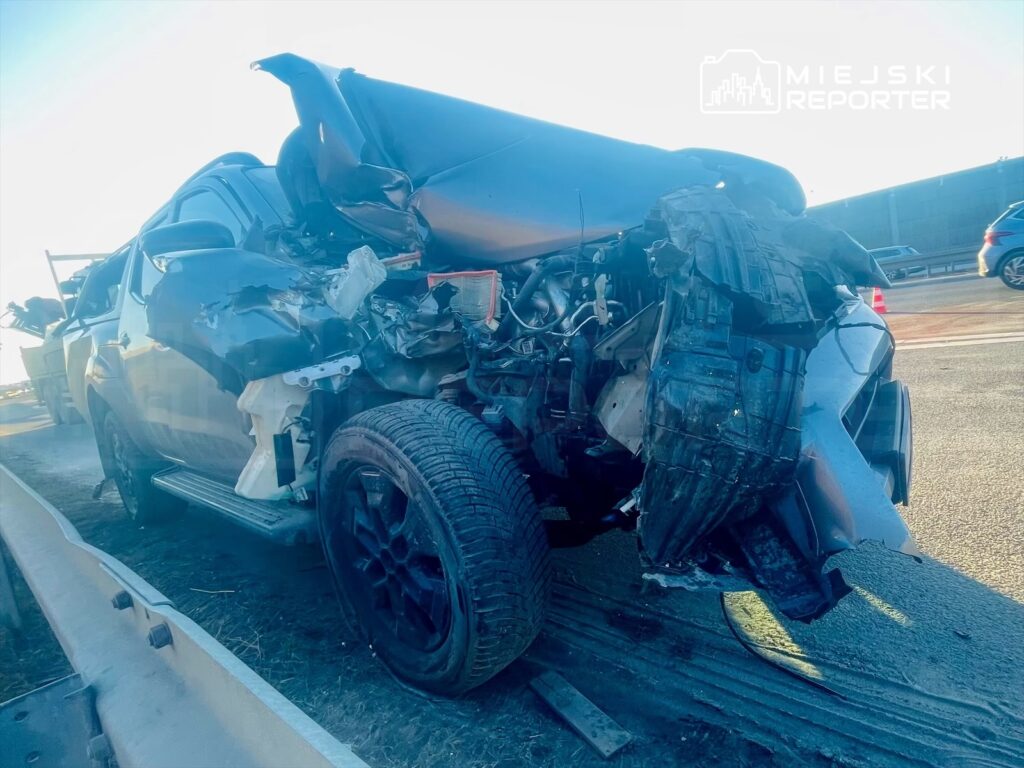 Uszkodzony samochód osobowy z rozdartą karoserią stoi na poboczu drogi, obok widać inne pojazdy.