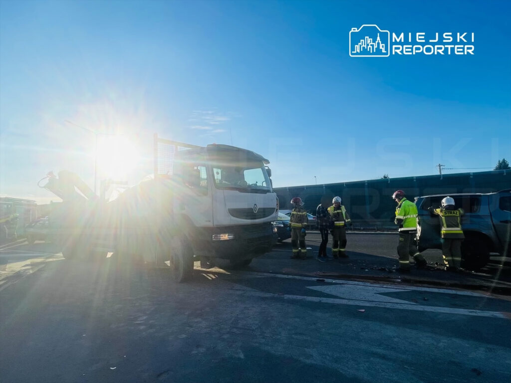 Służby ratunkowe rozmawiają z poszkodowanym obok rozbitego pojazdu na drodze, w tle widać wschodzące słońce.