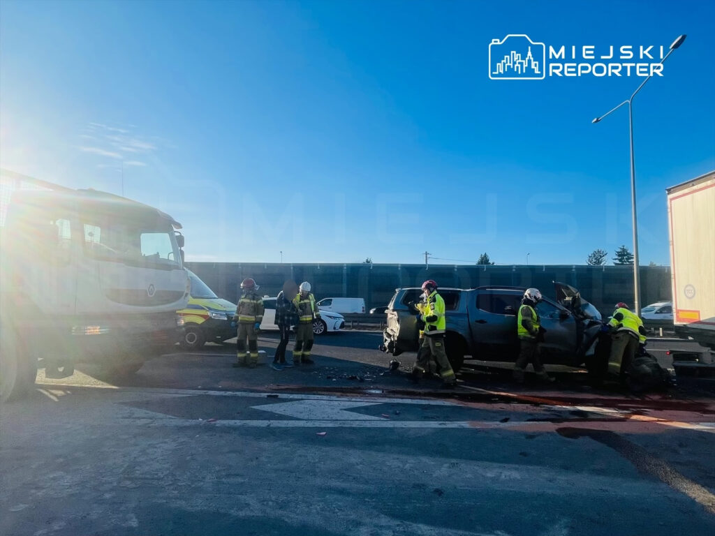 Ratownicy w kamizelkach odblaskowych pracują przy rozbitym samochodzie na drodze, obok stoi wóz strażacki.