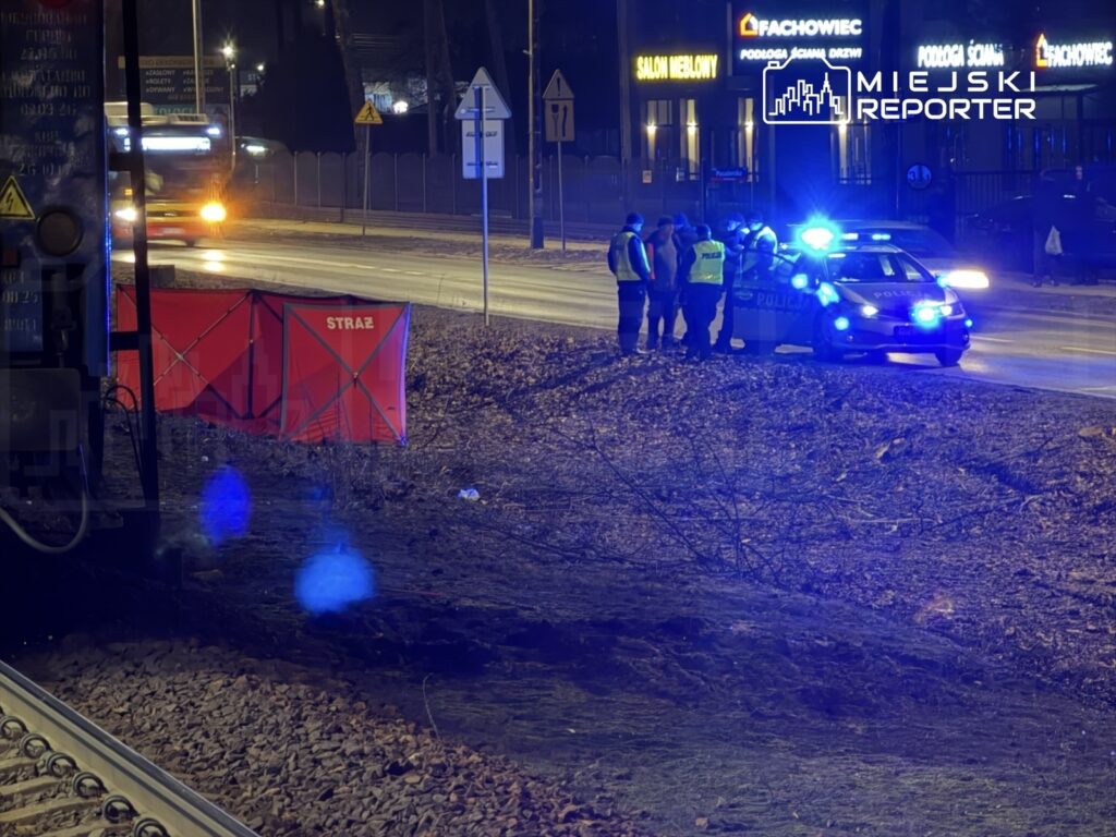 Grupa policjantów rozmawia obok radiowozu z włączonymi sygnałami świetlnymi na poboczu drogi nocą. W tle widać czerwony namiot z napisem "STRAŻ".