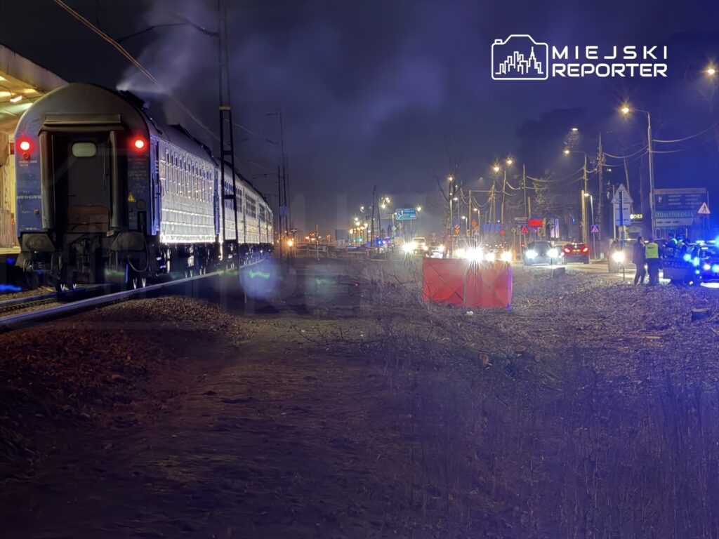 Pociąg towarowy zatrzymany przy peronie, w tle policyjne światła i ruch uliczny w nocy.