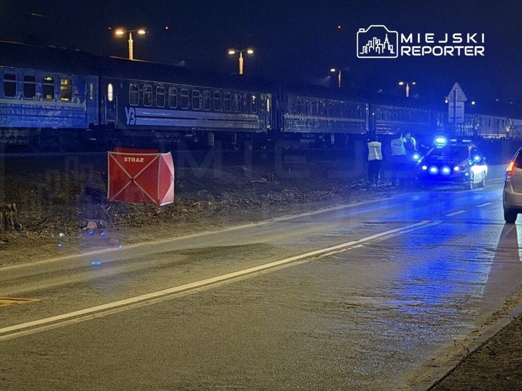 Grupa osób w odblaskowych kamizelkach rozmawia obok radiowozu z włączonymi sygnalizatorami przy torach kolejowych w nocy.