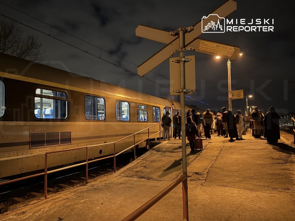 Grupa pasażerów czeka na peronie przy pociągu nocnym, oświetlonym lampami, w otoczeniu ciemnego nieba.