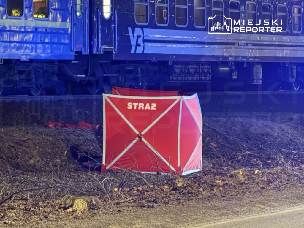 Czerwony namiot strażacki ustawiony obok torów kolejowych, w tle widoczny niebieski pociąg.