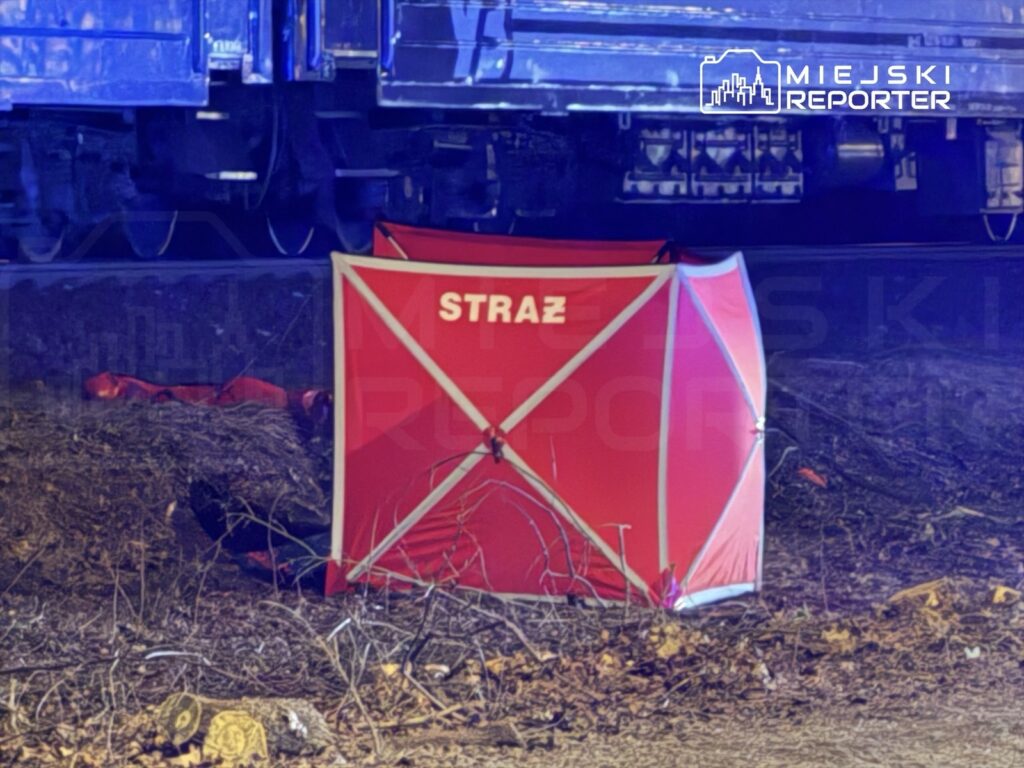 Czerwony namiot ochronny z napisem "STRAŻ" ustawiony obok torów kolejowych w lesie.