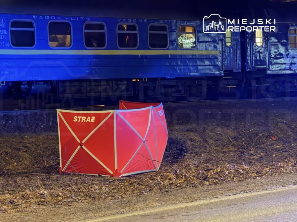 Czerwony namiot z napisem „STRAŻ” ustawiony obok torów kolejowych, w tle niebieski wagon pociągu.