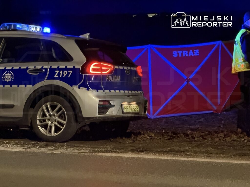 Radiowóz policyjny z włączonymi sygnałami świetlnymi stoi obok czerwonego namiotu z napisem "STRAŻ" w ciemnym otoczeniu.