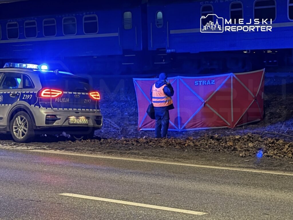 Policjant w kamizelce odblaskowej rozmawia z ratownikiem przy czerwonym namiocie straży obok torów kolejowych.