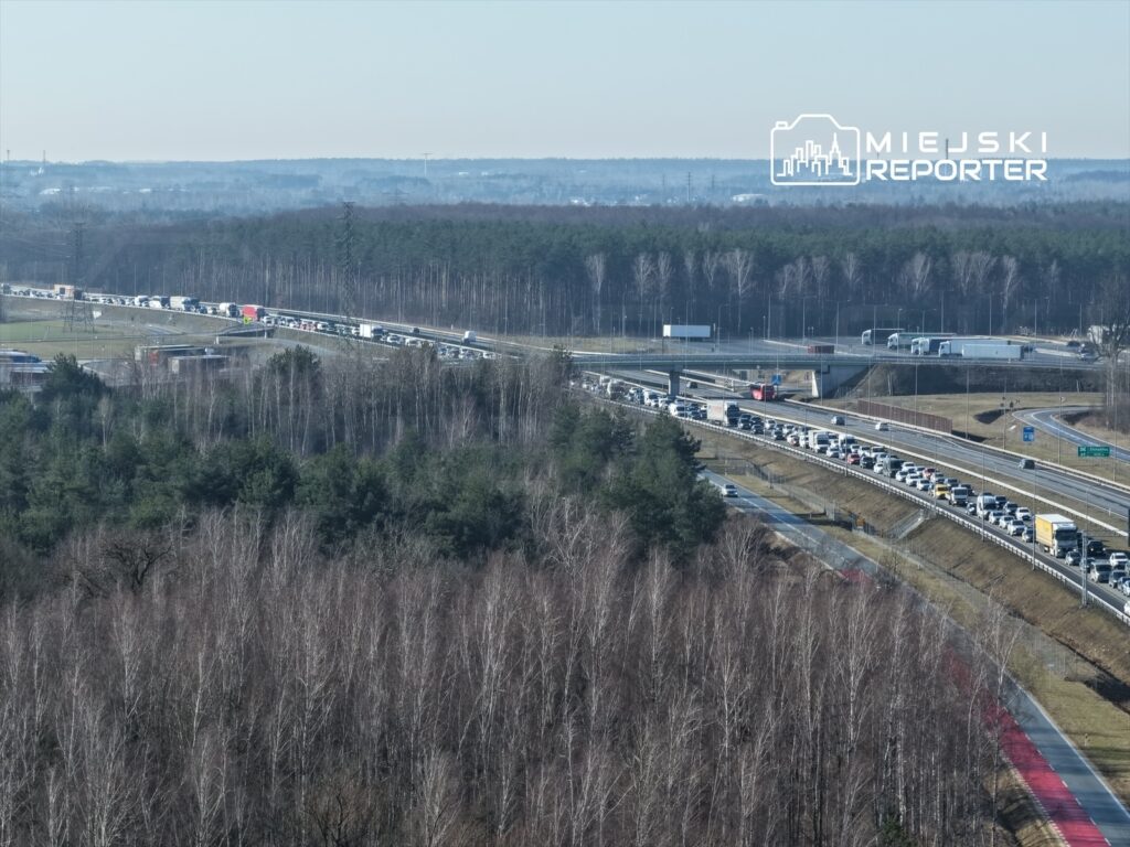 Długa kolumna pojazdów stojących w korku na drodze otoczonej lasem, z widocznymi zjazdami i ciężarówkami w tle.
