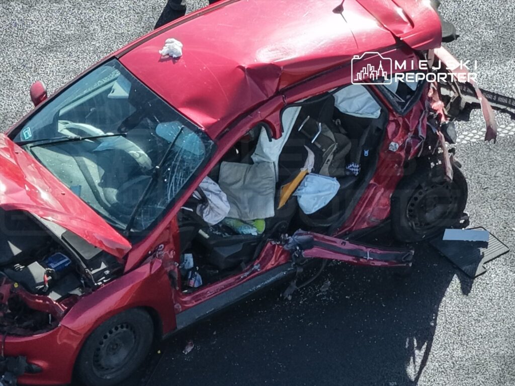 Czerwony samochód osobowy z poważnymi uszkodzeniami leży na drodze, z otwartymi drzwiami i widoczną bagażówką.