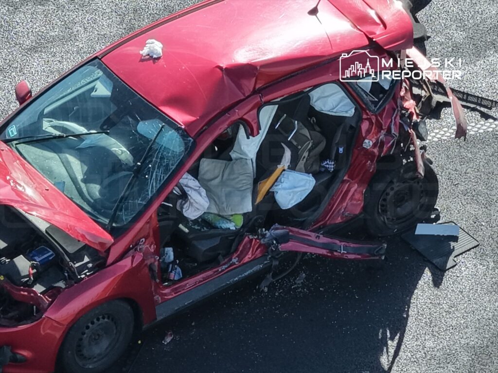 Czerwony samochód osobowy zniszczony w wyniku wypadku, z otwartymi drzwiami i porozrzucanymi przedmiotami w środku.