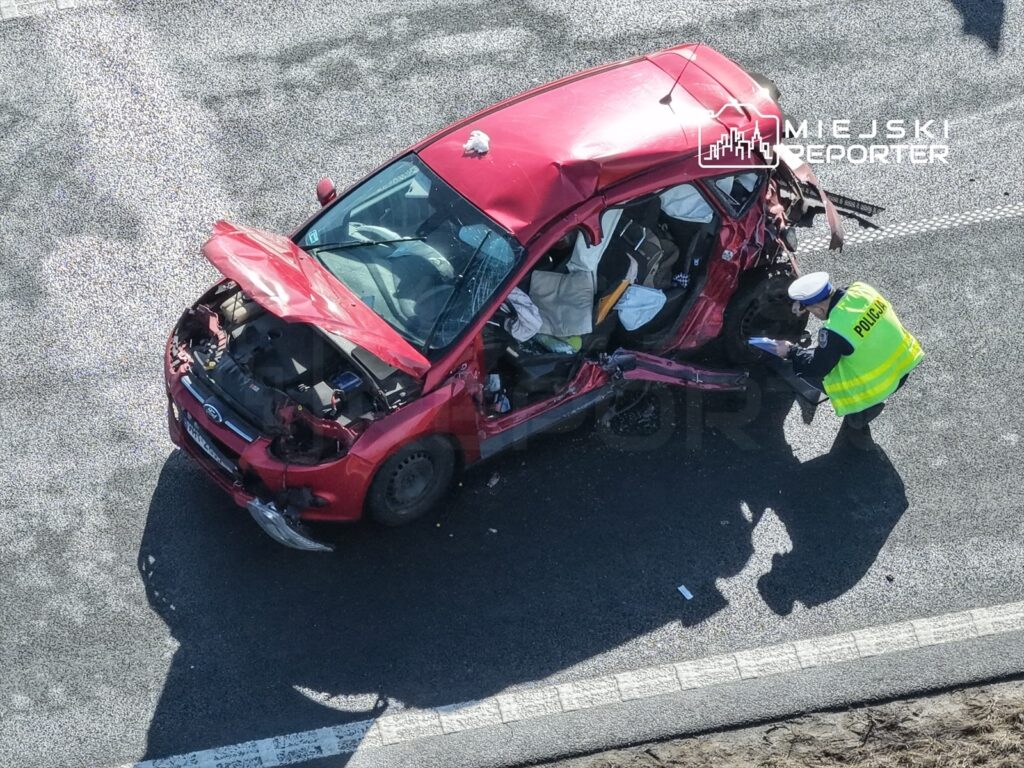 Policjant w kamizelce odblaskowej bada uszkodzony czerwony samochód osobowy na drodze.
