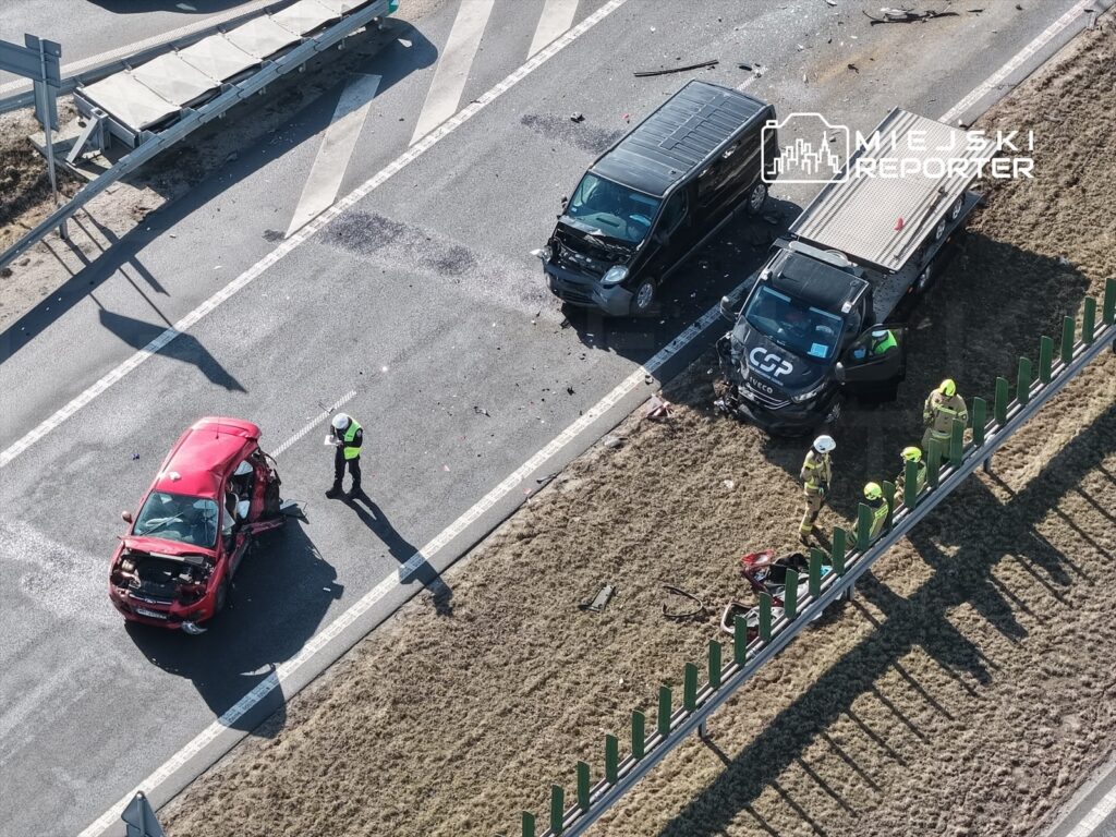 Czerwony samochód osobowy z uszkodzoną karoserią stoi na poboczu drogi, obok pracują strażacy i policjant w kamizelce odblaskowej.