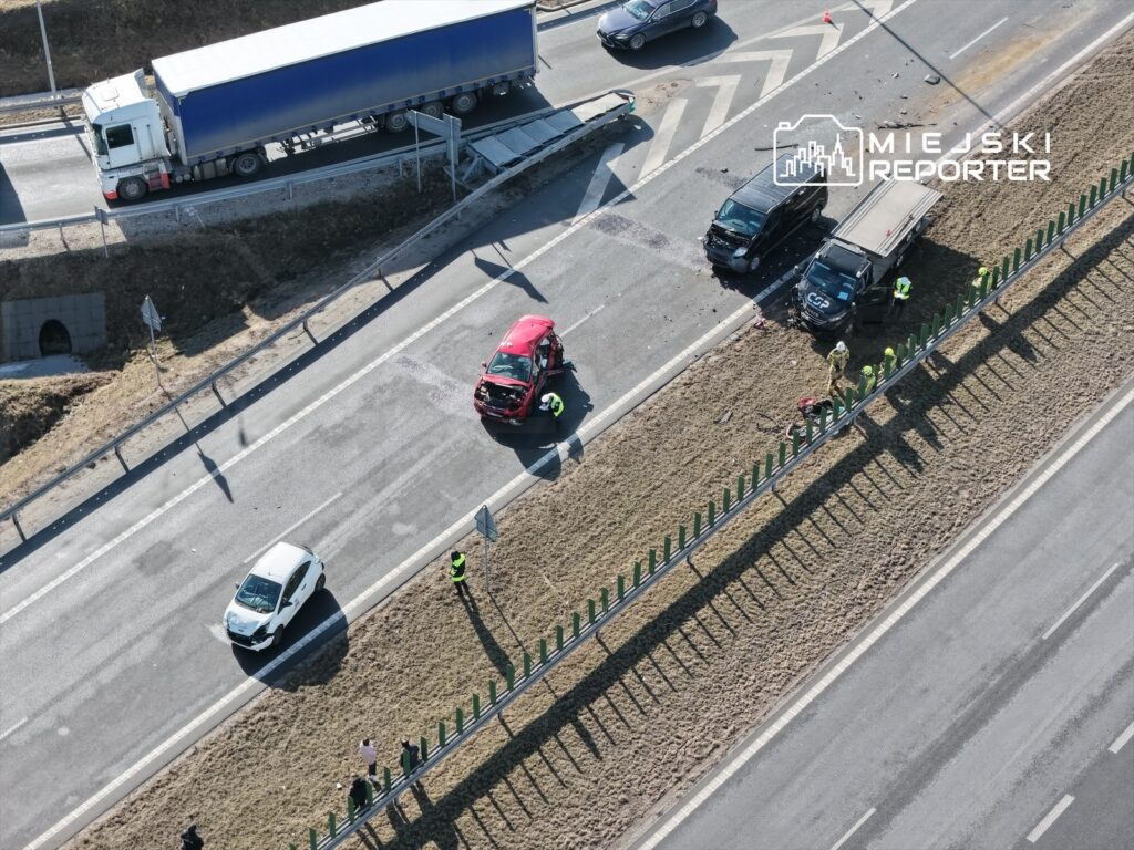 Czerwony samochód osobowy z uszkodzoną maską stoi na drodze, obok pracują strażacy i policjanci, a w tle widać ciężarówkę.