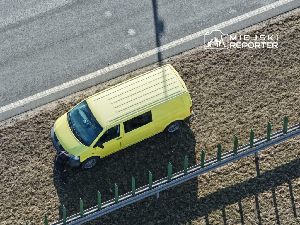 Mężczyzna w czarnej odzieży naprawia żółty samochód dostawczy na poboczu drogi, obok metalowego ogrodzenia.