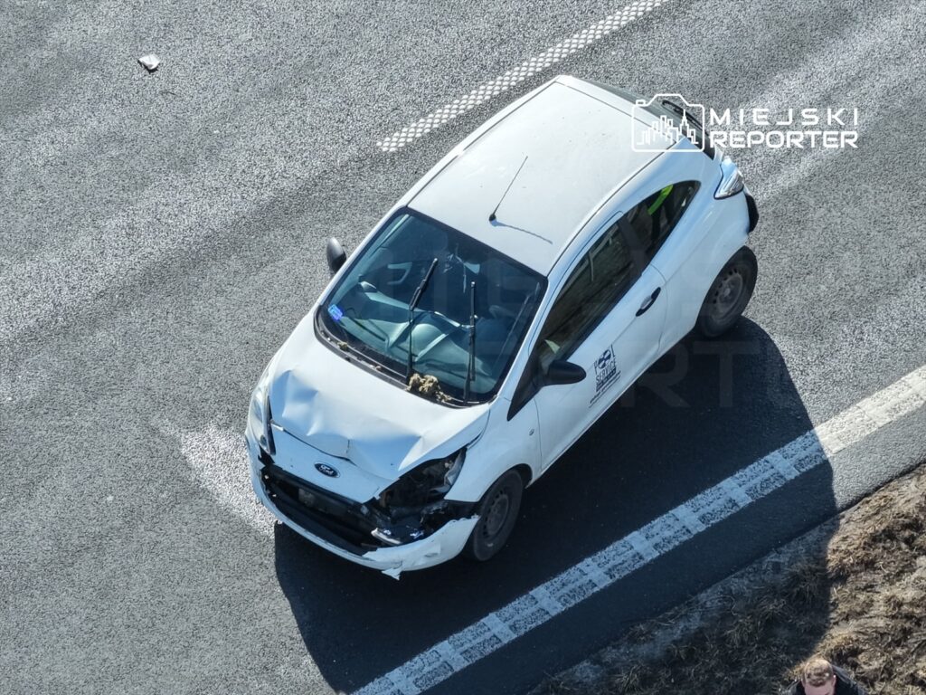 Uszkodzony biały samochód osobowy zaparkowany na drodze, z widocznymi uszkodzeniami przodu.