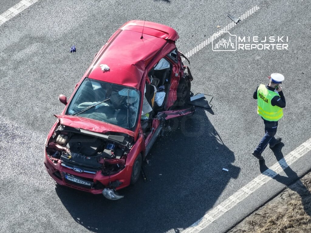 Czerwony samochód osobowy z uszkodzoną karoserią stoi na drodze, obok policjant w kamizelce odblaskowej rozmawia przez telefon.
