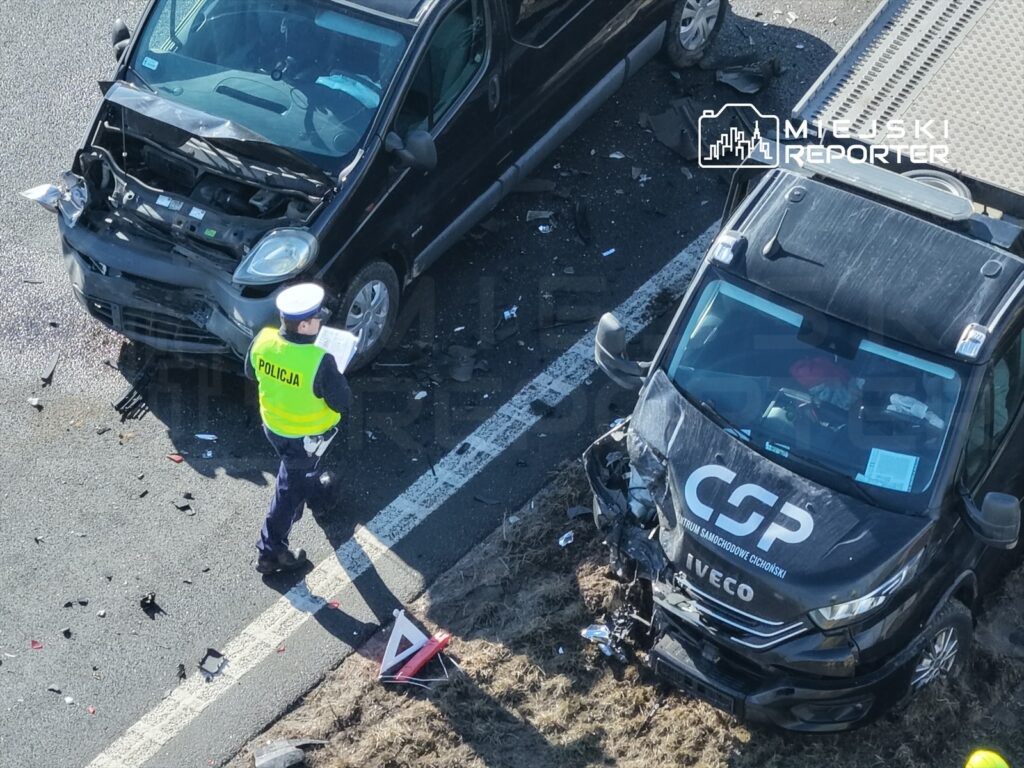 Policjant w kamizelce odblaskowej sporządza notatki na miejscu wypadku drogowego, obok uszkodzonych pojazdów.