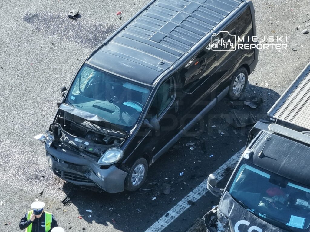 Czarny dostawczy samochód z uszkodzoną maską stoi na jezdni, obok policjant w kamizelce odblaskowej prowadzi czynności.
