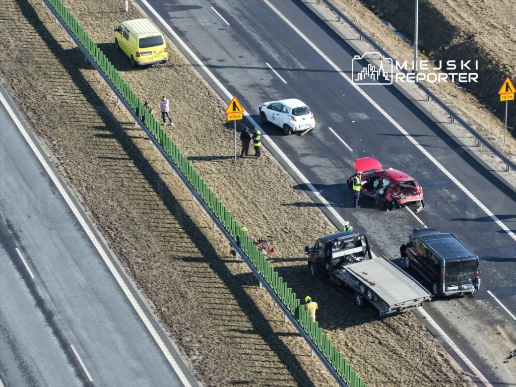 W miejscu wypadku drogowego, ratownicy pracują nad uszkodzonym czerwonym samochodem, obok stoi laweta.