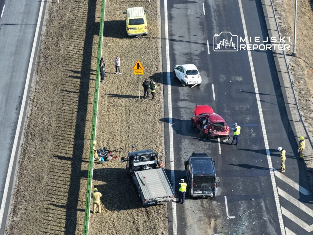 Na drodze leży rozbity czerwony samochód, obok niego czarny pojazd oraz strażacy i policjanci zabezpieczający miejsce wypadku.