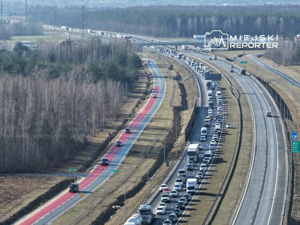 Ruch samochodowy na autostradzie z długim zatoriem, obok widoczna droga rowerowa w otoczeniu drzew.